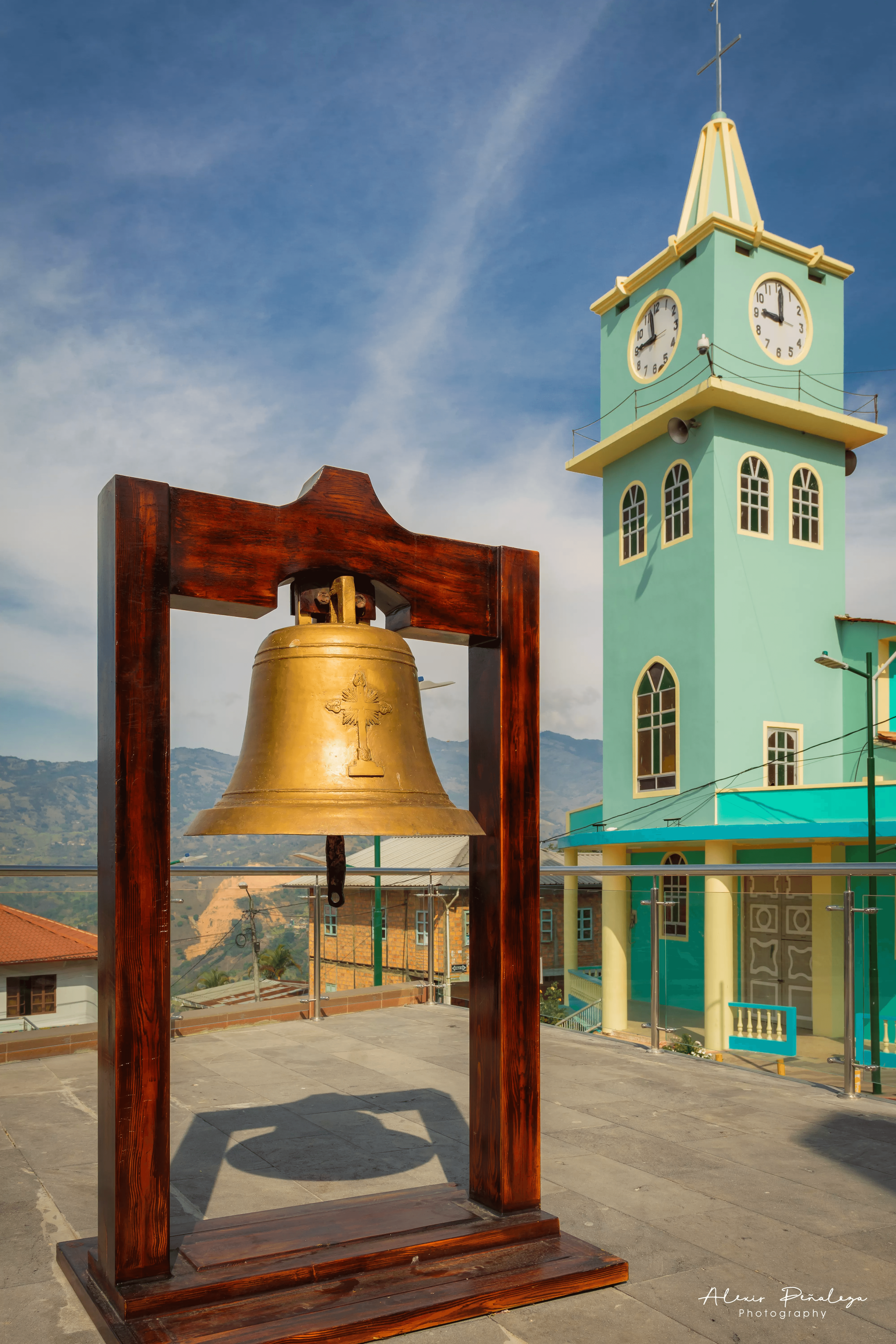Campana de bronce en soporte de madera con la torre de la iglesia al fondo