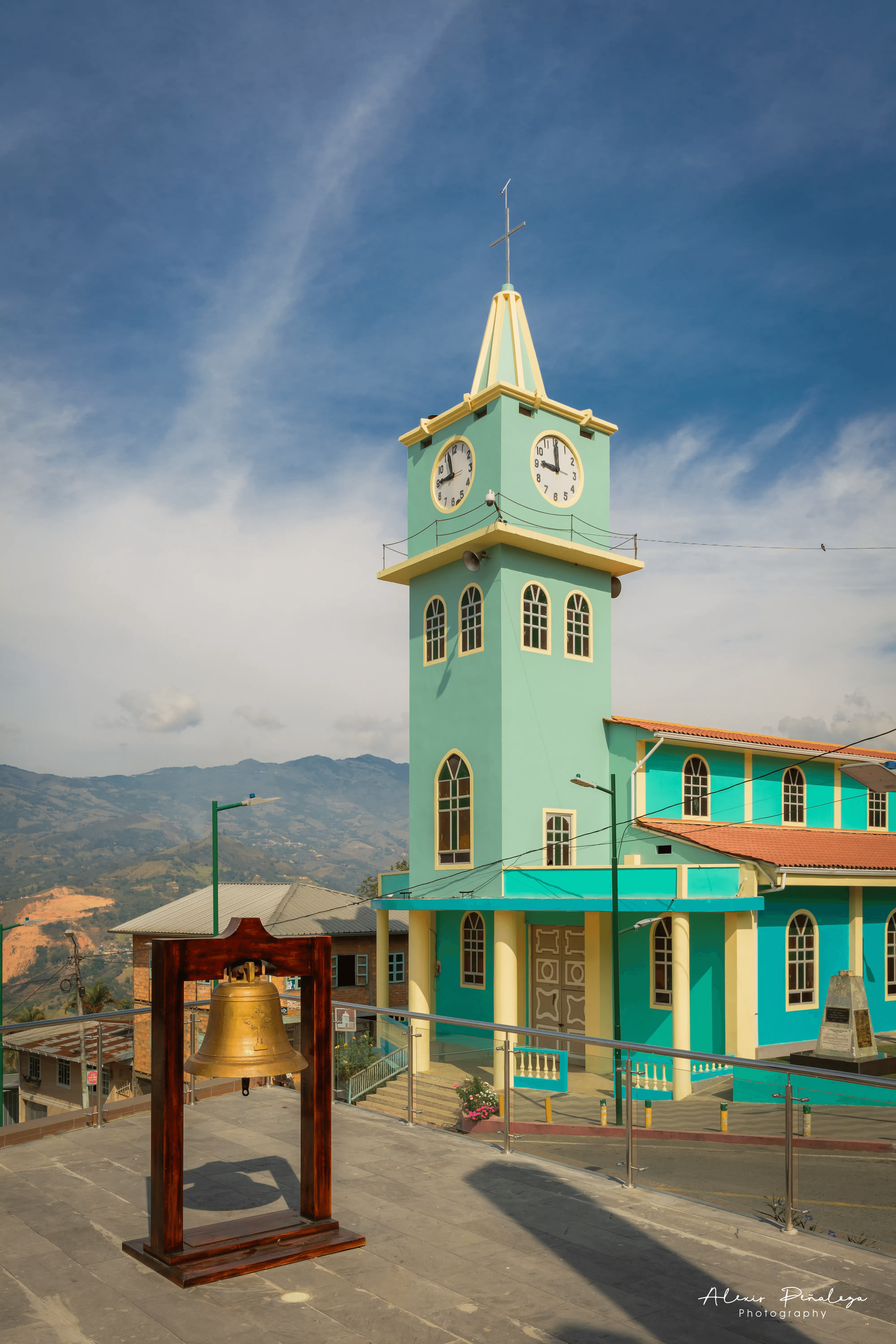 Campana histórica de bronce en primer plano frente a la iglesia turquesa