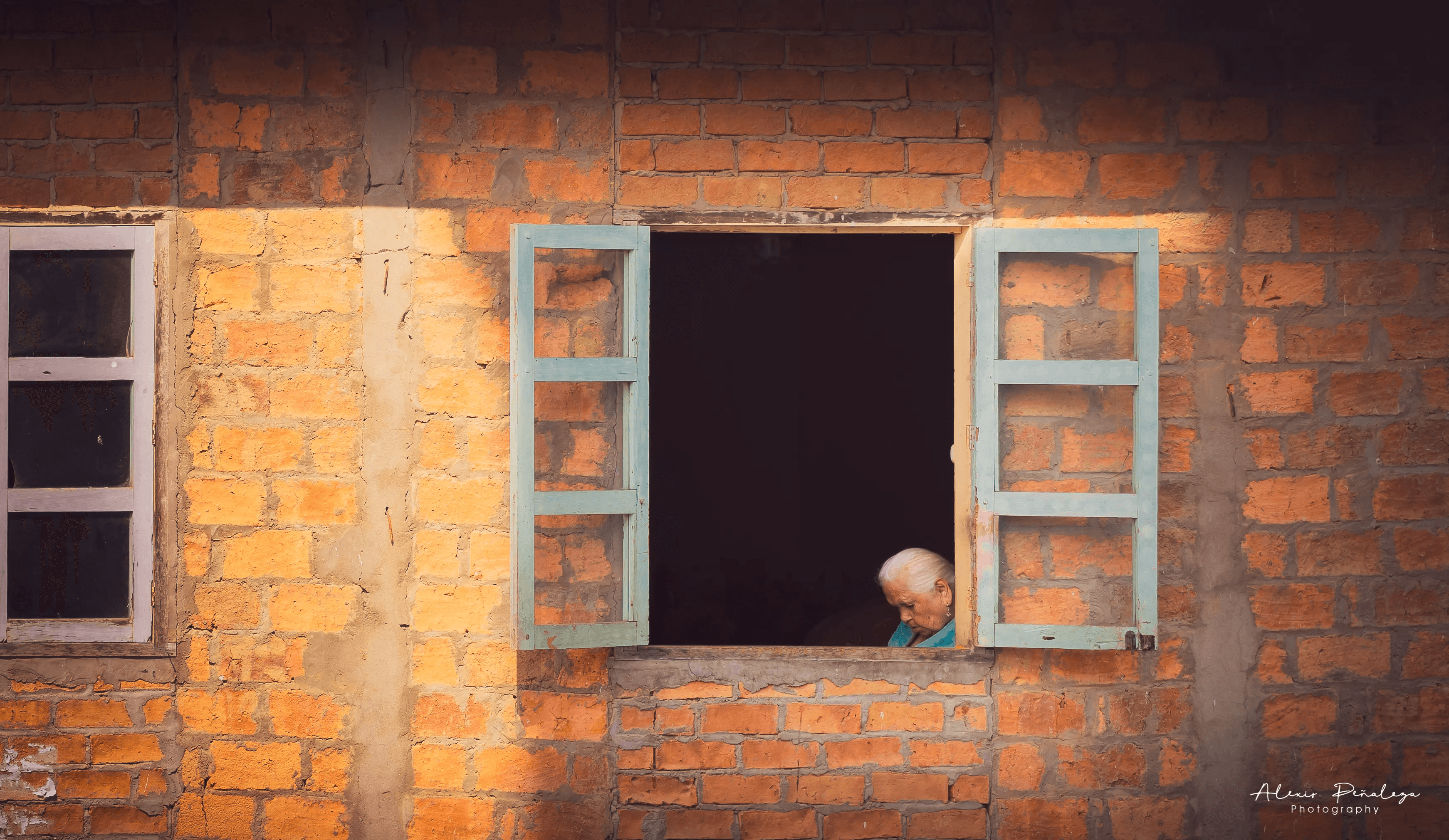 Anciana de cabello blanco asomándose apenas en la ventana de una pared cálida de ladrillos