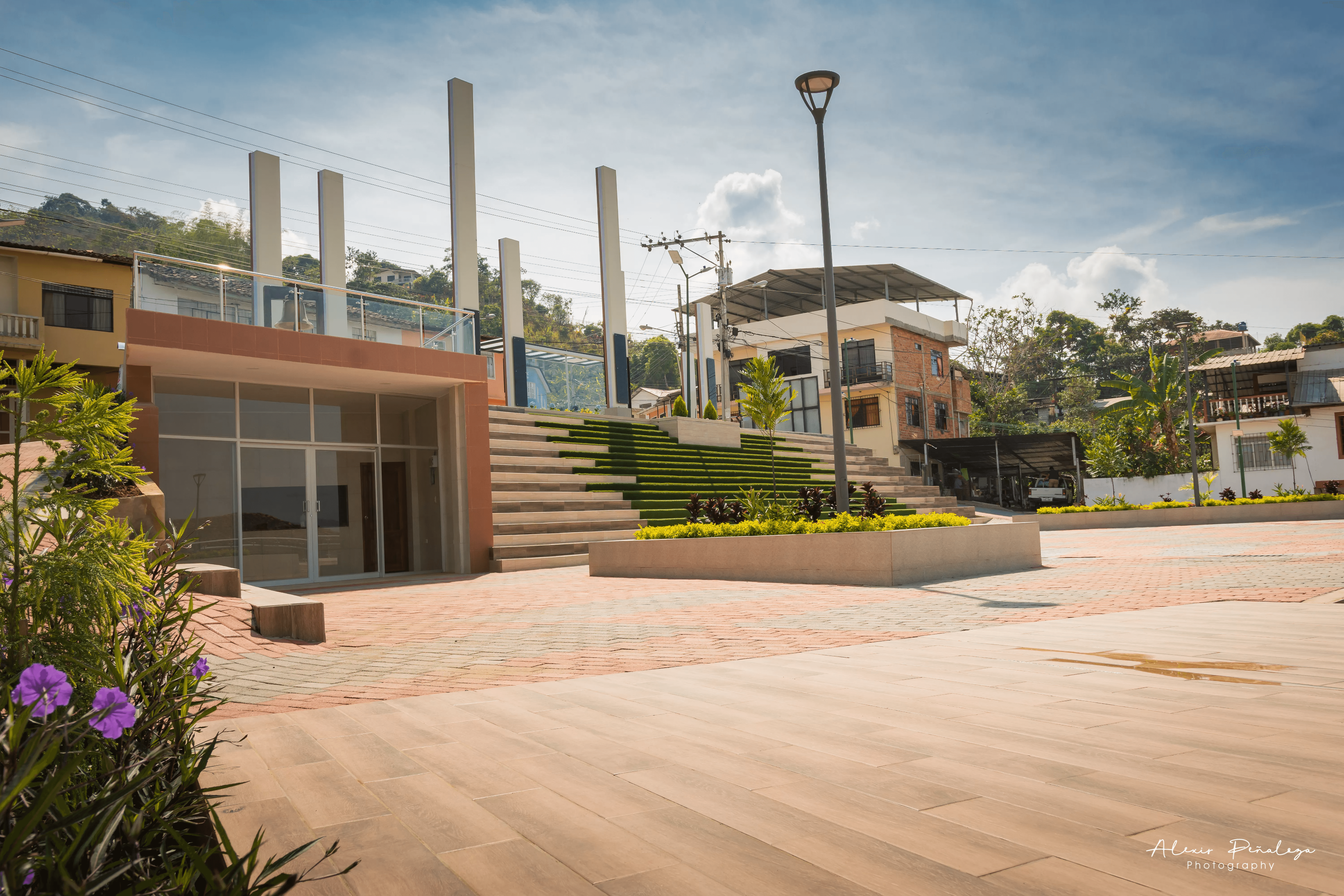 Amplia plaza peatonal esquinera con graderío verdoso y un edificio moderno con balcón
